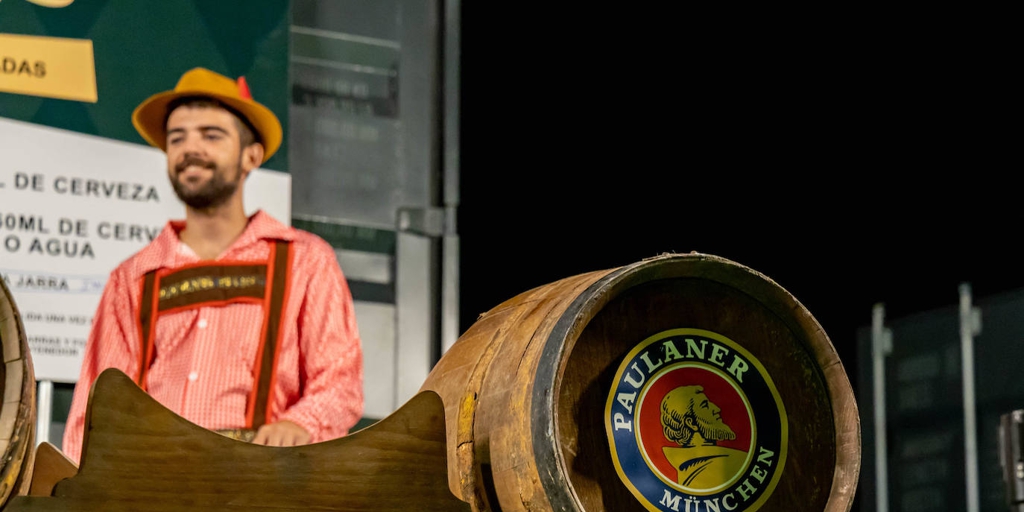 Un hombre vestido de manera típica frente a un barril de cerveza de madera con la marca Paulaner